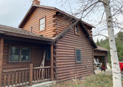 A two-story log cabin with a covered porch and bare trees nearby.